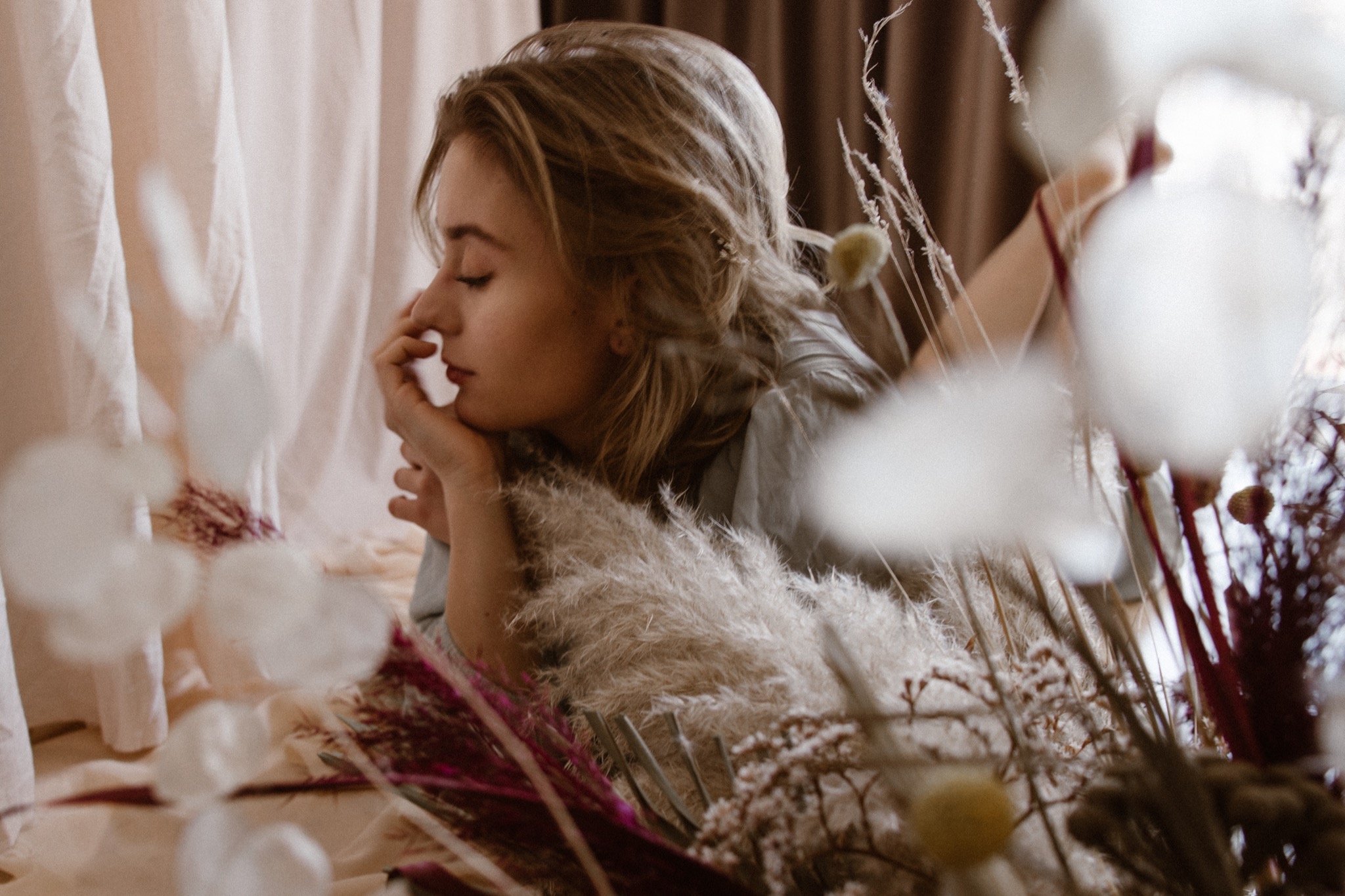 Kobieta z zamkniętymi oczami, podpierająca brodę dłonią, otoczona dekoracjami z suszonych kwiatów w odcieniach beżu i bordo na tle jasnych drapowanych tkanin.