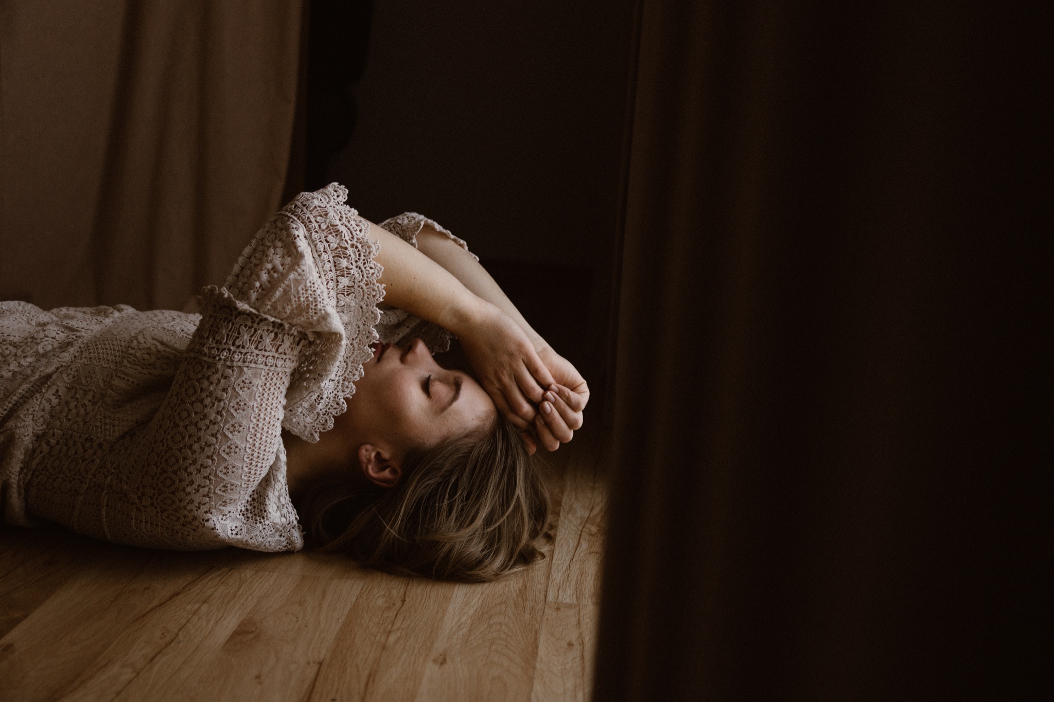 Kobieta w koronkowej sukience leży na drewnianej podłodze, dłonie splecione nad głową, oczy zamknięte, w tle zasłony.