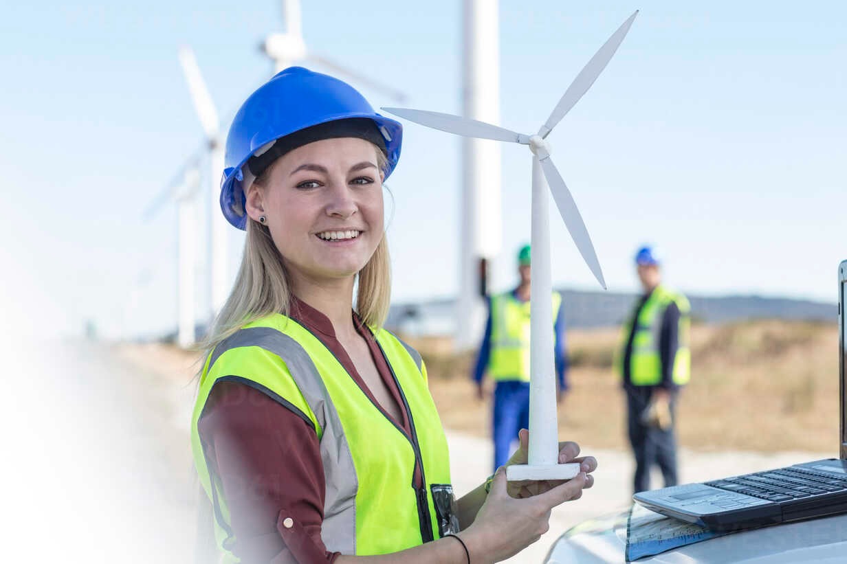 Uśmiechnięta kobieta w kasku i kamizelce odblaskowej trzyma model turbiny wiatrowej na tle farmy wiatrowej i laptopa.