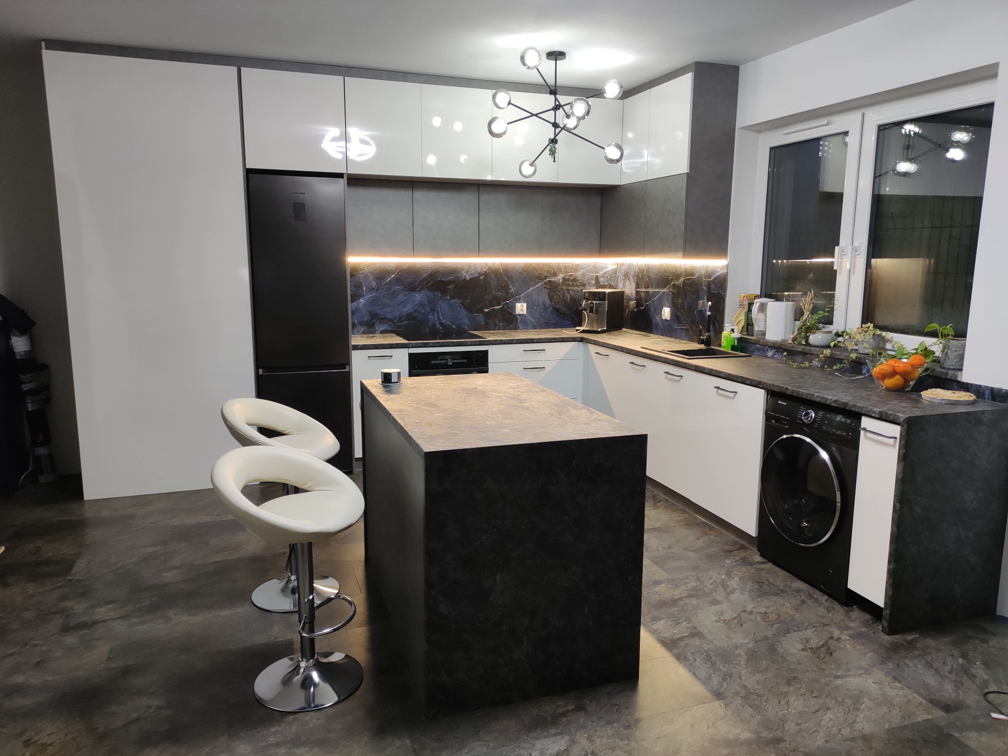 Nowoczesna kuchnia z wyspą wykończoną blatem z ciemnego kamienia, oświetleniem LED pod szafkami i dekoracyjną lampą wiszącą.