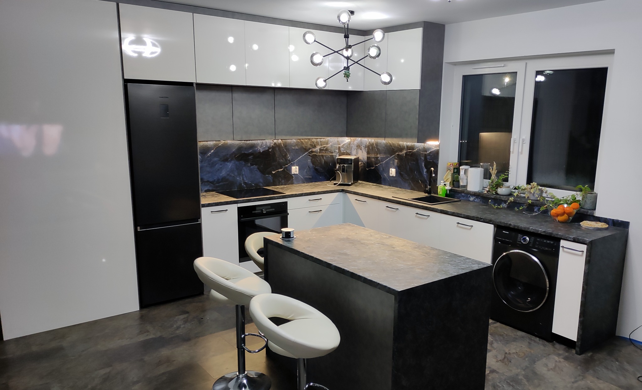 Nowoczesna kuchnia z wyspą, białymi szafkami i czarnym blatem imitującym marmur, oświetlona lampą sufitową w stylu atomu, z trzema białymi hokerami przy wyspie.