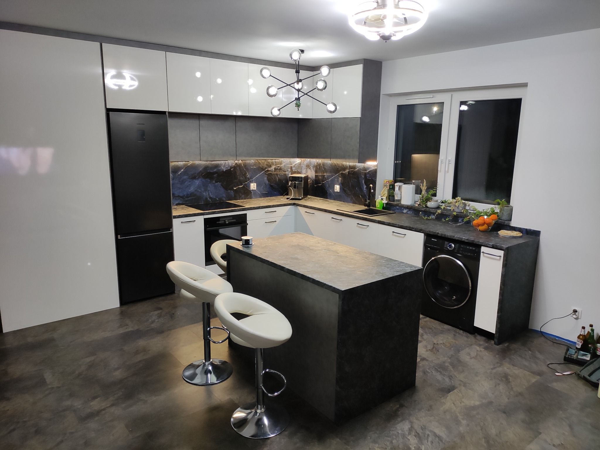 Nowoczesna kuchnia z wyspą, białymi szafkami górnymi z połyskiem, szarymi szafkami dolnymi i blatem z ciemnego kamienia z marmurkowym wzorem. Na wyspie stoją dwa białe hokery z chromowanymi nogami.
