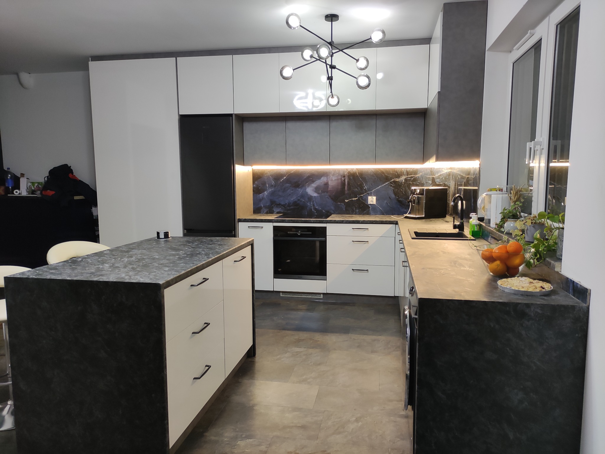 Nowoczesna kuchnia z białymi szafkami i ciemnoszarym blatem z efektem marmuru, oświetleniem LED pod szafkami wiszącymi, wyspą kuchenną i designerską lampą sufitową.