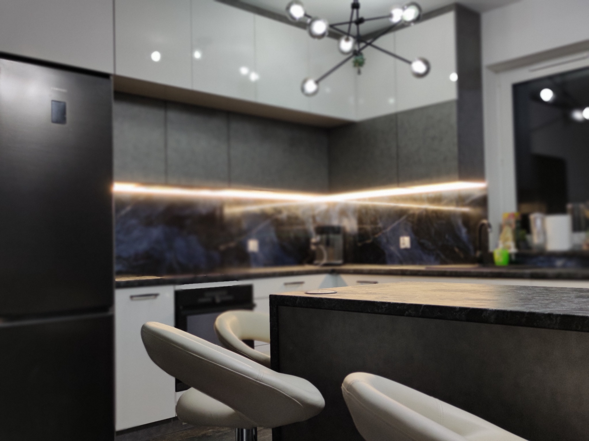 Nowoczesna kuchnia z wyspą, blatem z ciemnego kamienia i podświetleniem LED pod szafkami, widoczne białe hokery i czarna lodówka.