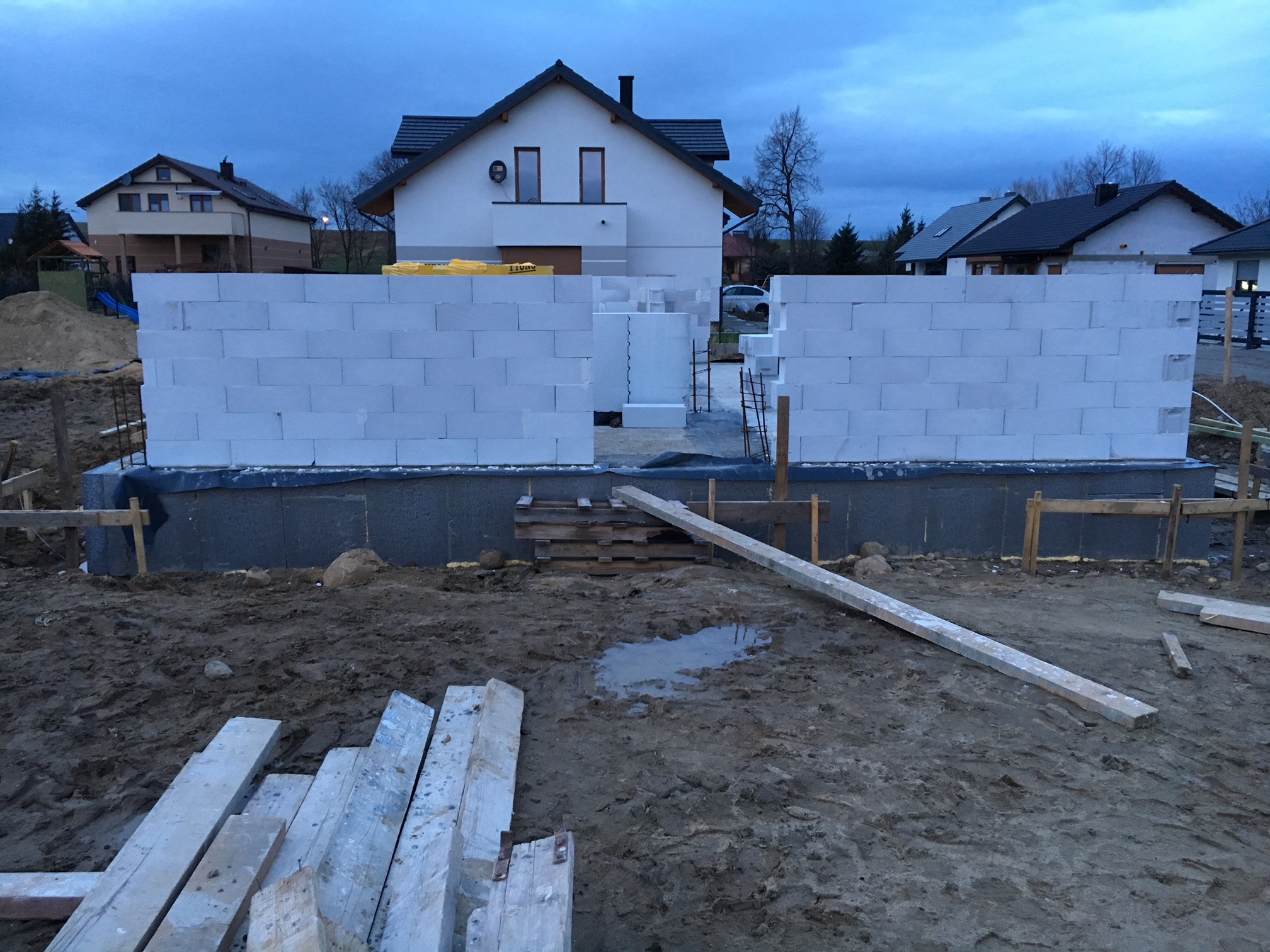 Wznoszenie ścian parteru z bloczków betonu komórkowego na fundamencie, widoczne drewniane szalunki i częściowo wybudowane ściany domu w tle, pochmurne niebo.