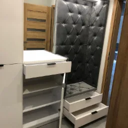 Biała szafa na wymiar z otwartymi szufladami i półkami, wbudowane tapicerowane siedzisko z pikowanym oparciem w kolorze szarym.