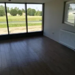 Wnętrze pokoju z nową, ciemną podłogą panelową ułożoną wzdłuż okna, grzejnik pod oknem, widok na zielony krajobraz za oknem.