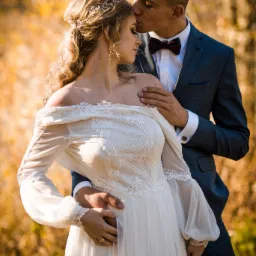 Elegancka para młoda w jesiennej scenerii: panna młoda w białej sukni z koronkowymi detalami i ozdobą we włosach, pan młody w granatowym garniturze z bordową muchą, czule całujący pannę młodą w czoło.