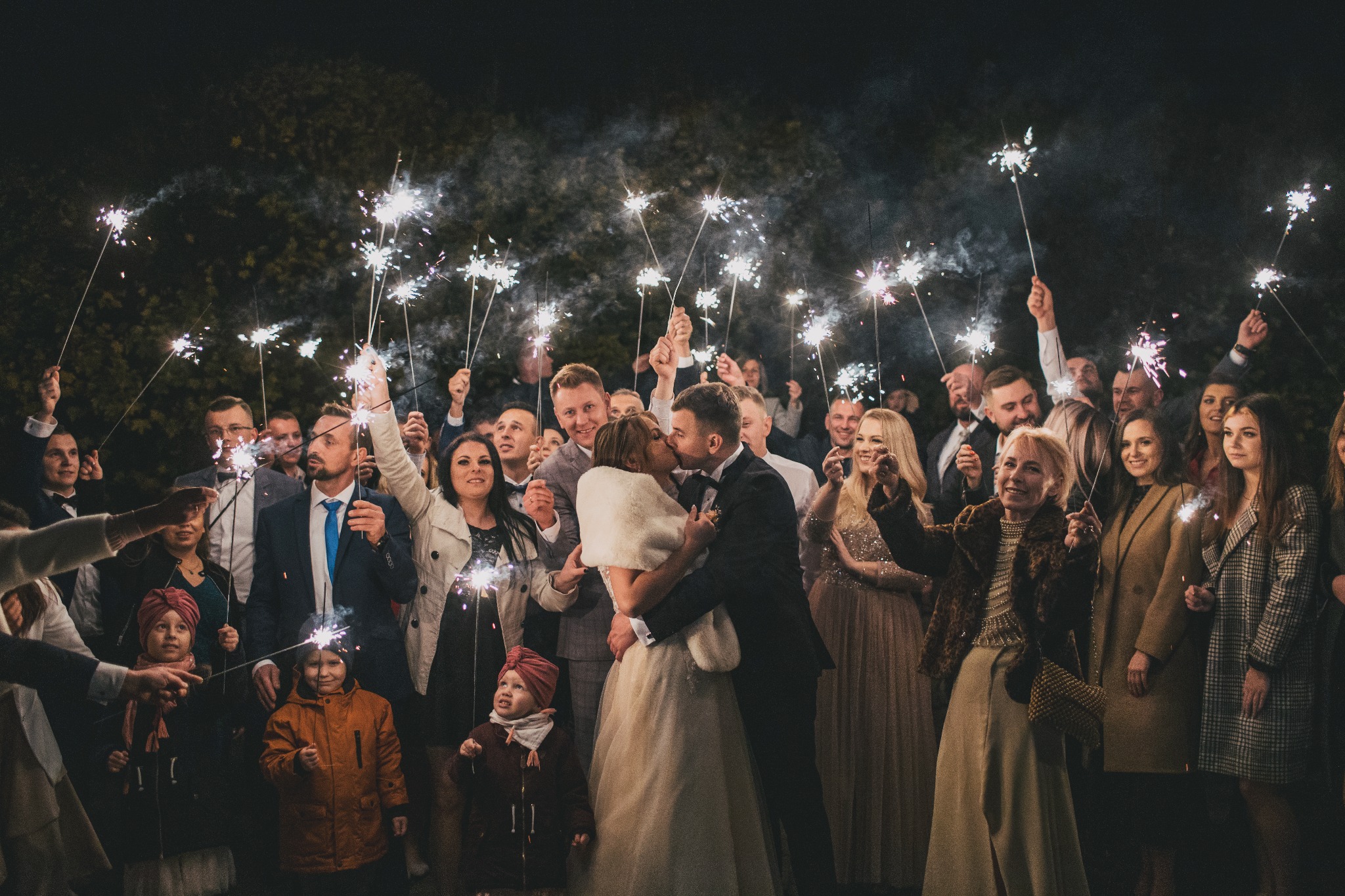 Nowożeńcy całują się, otoczeni gośćmi weselnymi trzymającymi zapalone zimne ognie, w tle ciemne drzewa.