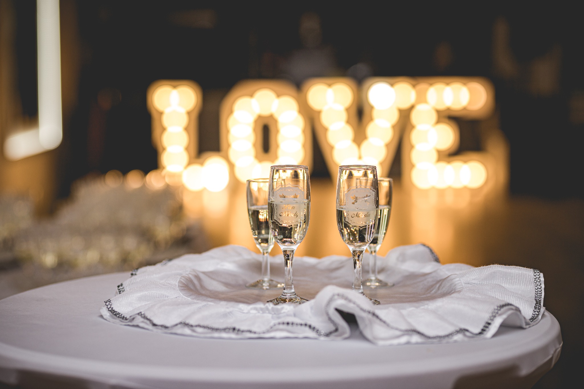 Cztery kieliszki do szampana z grawerem 'Żon Młody' i 'Żona Młoda' stojące na okrągłym stole przykrytym białym obrusem z koronkowym brzegiem, w tle napis LOVE z podświetlanych żarówek.