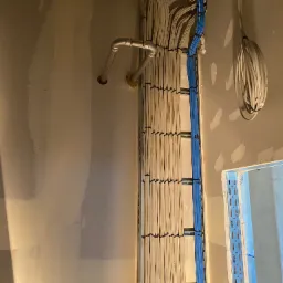 Ściana w trakcie remontu z równolegle ułożonymi kablami elektrycznymi w korytkach, częściowo zamaskowanymi, oraz rurą instalacyjną w rogu pomieszczenia.