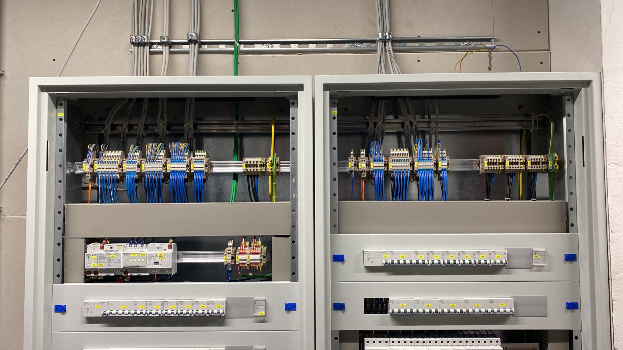 Dwa otwarte panele elektryczne z licznymi niebieskimi przewodami, listwami zaciskowymi, bezpiecznikami i szynami DIN, zamontowane na szarej ścianie.