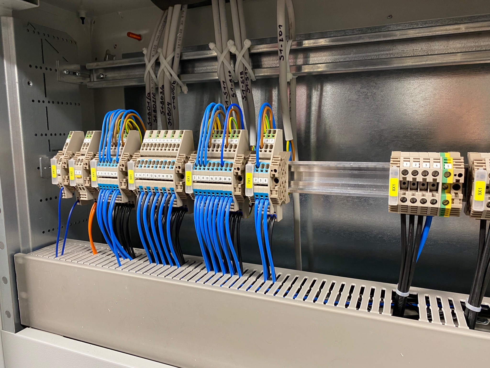 Rozdzielnica elektryczna z podłączonymi przewodami oznaczonymi etykietami, widoczne listwy zaciskowe z numeracją.