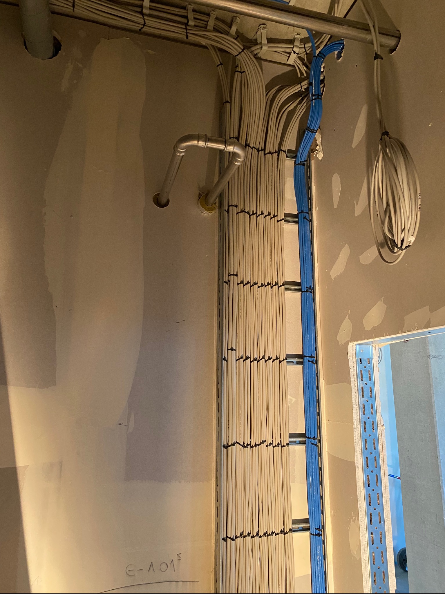 Ściana w trakcie remontu z równolegle ułożonymi kablami elektrycznymi w korytkach, częściowo zamaskowanymi, oraz rurą instalacyjną w rogu pomieszczenia.