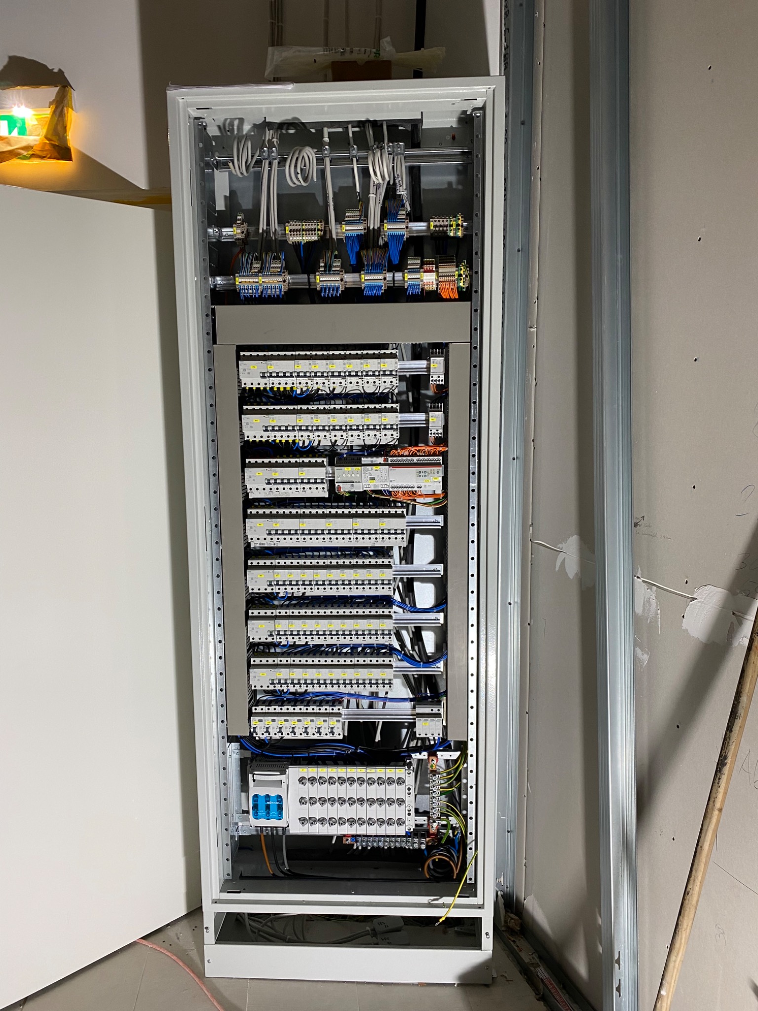 Otwarta szafa elektryczna z widocznymi bezpiecznikami, przewodami i elementami sterującymi, zamontowana w surowym wnętrzu z widocznymi profilami metalowymi na ścianie.