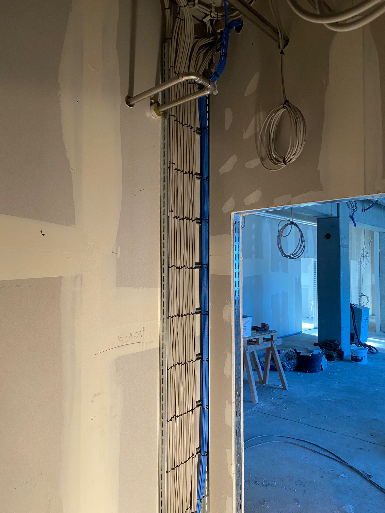 Ściana w trakcie wykończenia z pionowo ułożonymi kablami w korytkach, widoczne również kable u góry i w tle, surowy stan wykończenia wnętrza.