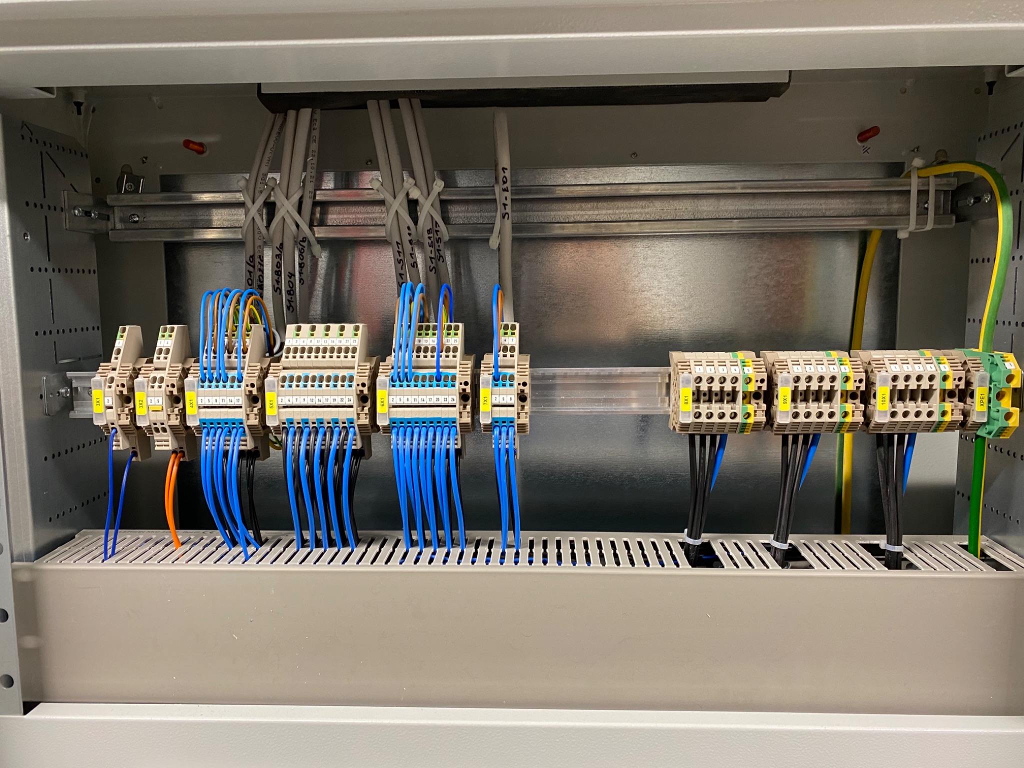 Rozdzielnica elektryczna w szarej obudowie z widocznymi listwami zaciskowymi, przewodami oznaczonymi numerami i różnymi kolorami, uporządkowanymi w wiązki, z przewodem uziemiającym w kolorze...