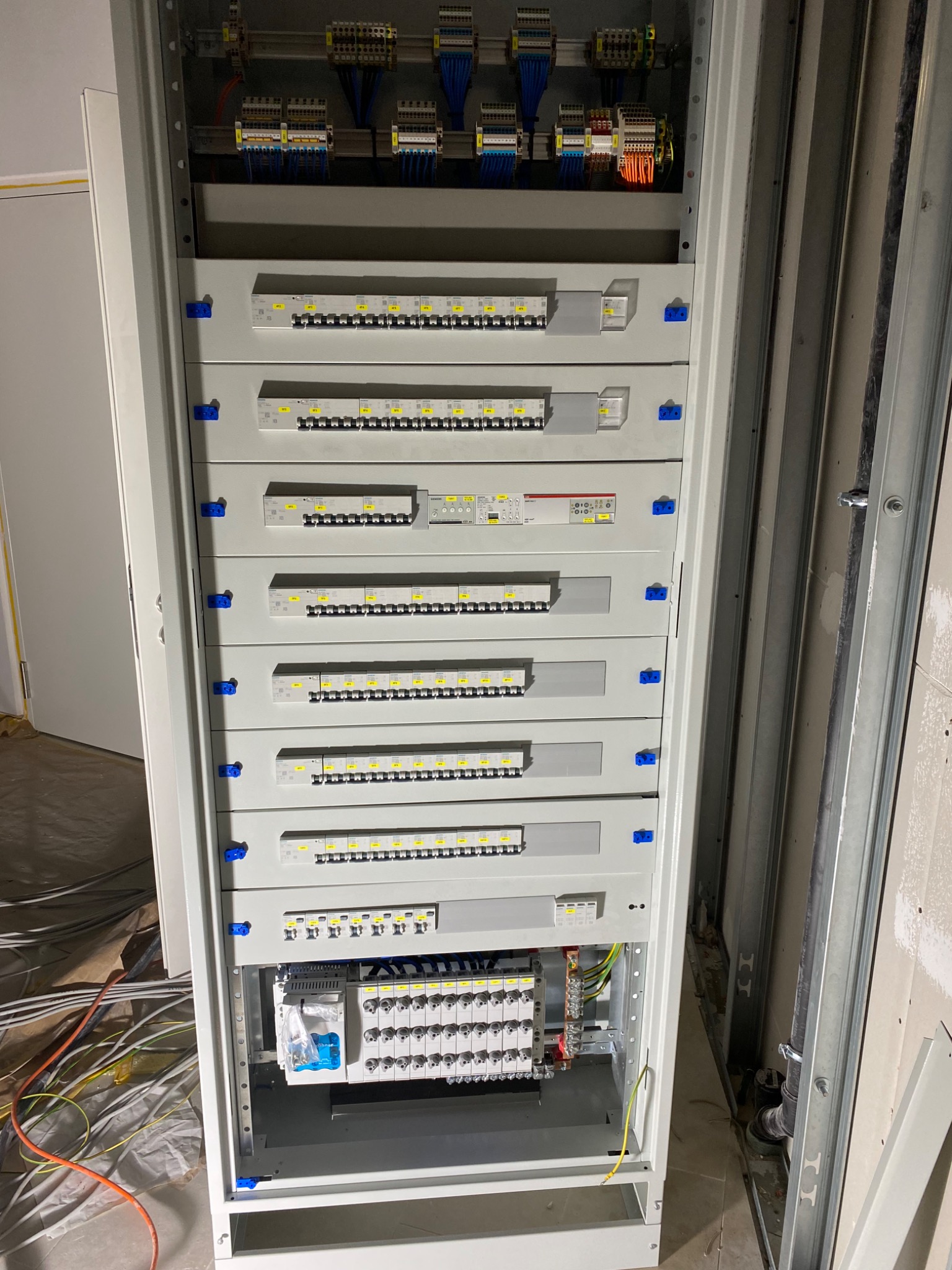 Rozdzielnica elektryczna z widocznymi bezpiecznikami, stycznikami i przewodami, w trakcie montażu w surowym wnętrzu. Na podłodze widoczne kable i folia ochronna.