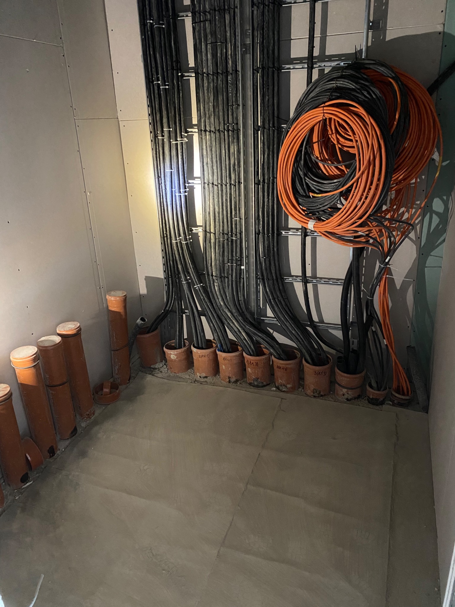 Układanie przewodów elektrycznych w rurach ochronnych w nowym budynku, widoczne kable czarne i pomarańczowe oraz mocowania do ściany.