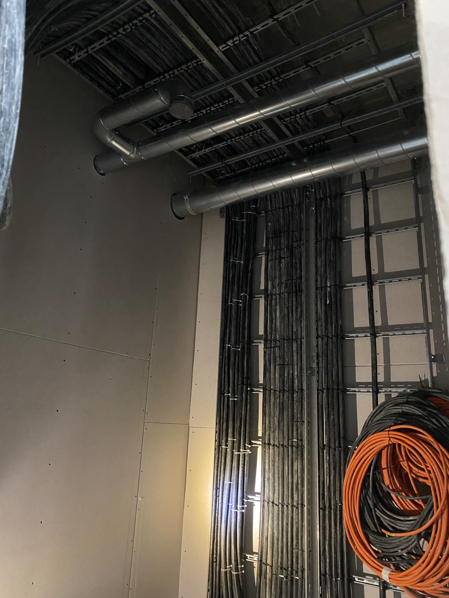 Wiązki kabli elektrycznych, czarnych i pomarańczowych, prowadzone w korytach pod sufitem w surowym stanie, widoczne rury wentylacyjne i metalowe drabinki kablowe.