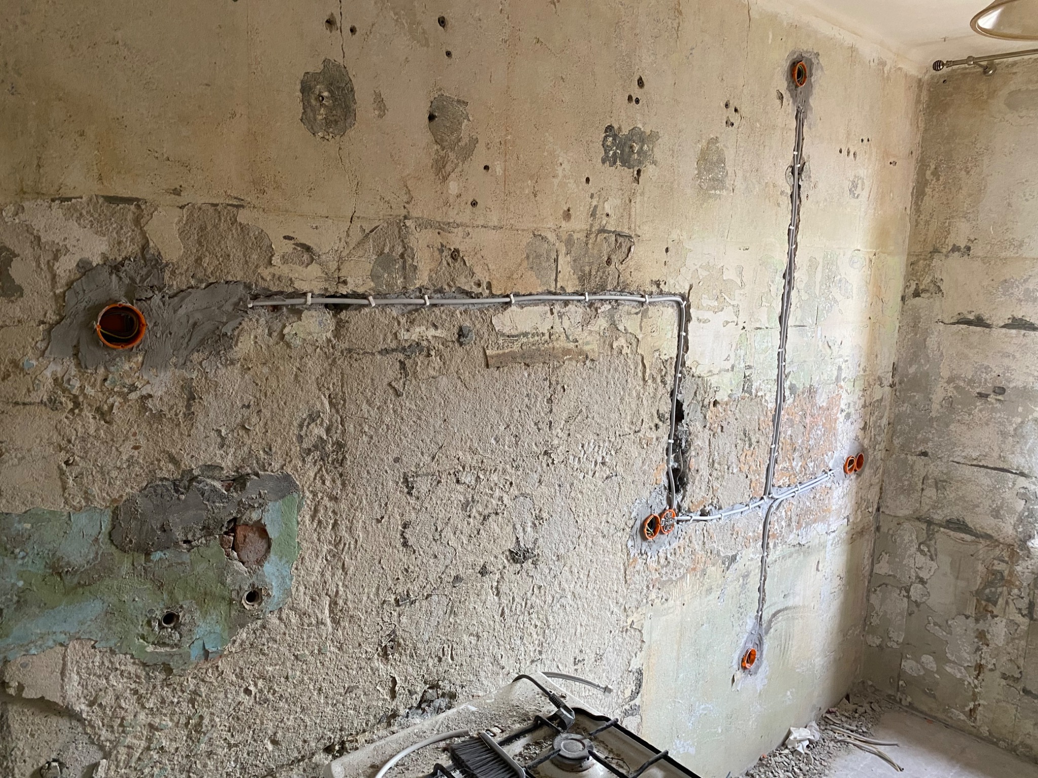 Ściana w trakcie remontu z widocznymi przewodami elektrycznymi w peszlach i pomarańczowymi puszkami instalacyjnymi, fragment kuchenki gazowej na dole kadru, surowy stan wykończenia.