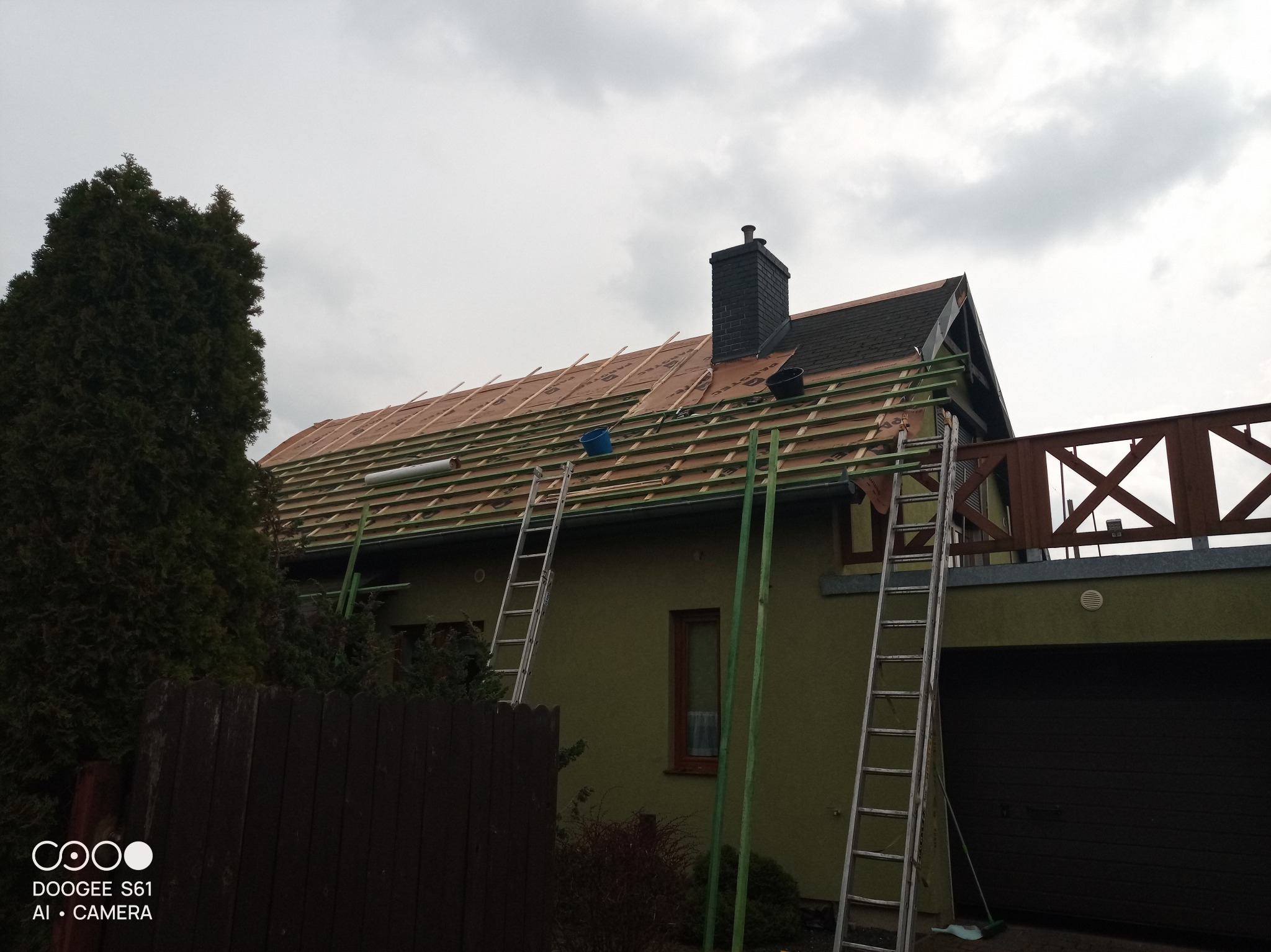 Montaż łat na dachu domu jednorodzinnego pod nową dachówkę, widoczne dwie drabiny opierające się o ścianę i dach, niebieskie wiaderko na dachu, pochmurne niebo.