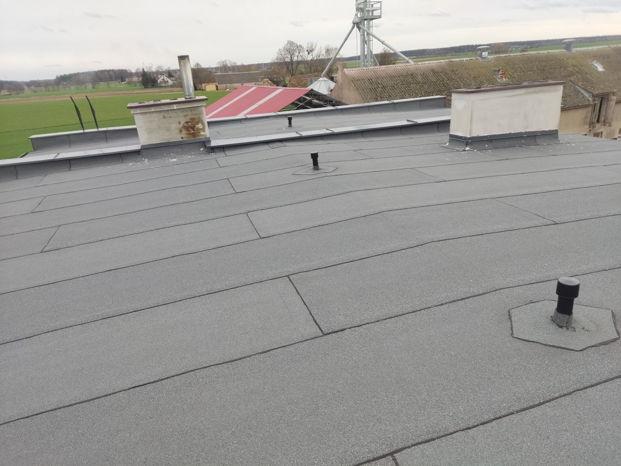 Szary, płaski dach pokryty papą termozgrzewalną z kominami i odpowietrznikami na tle krajobrazu wiejskiego z budynkami gospodarczymi.
