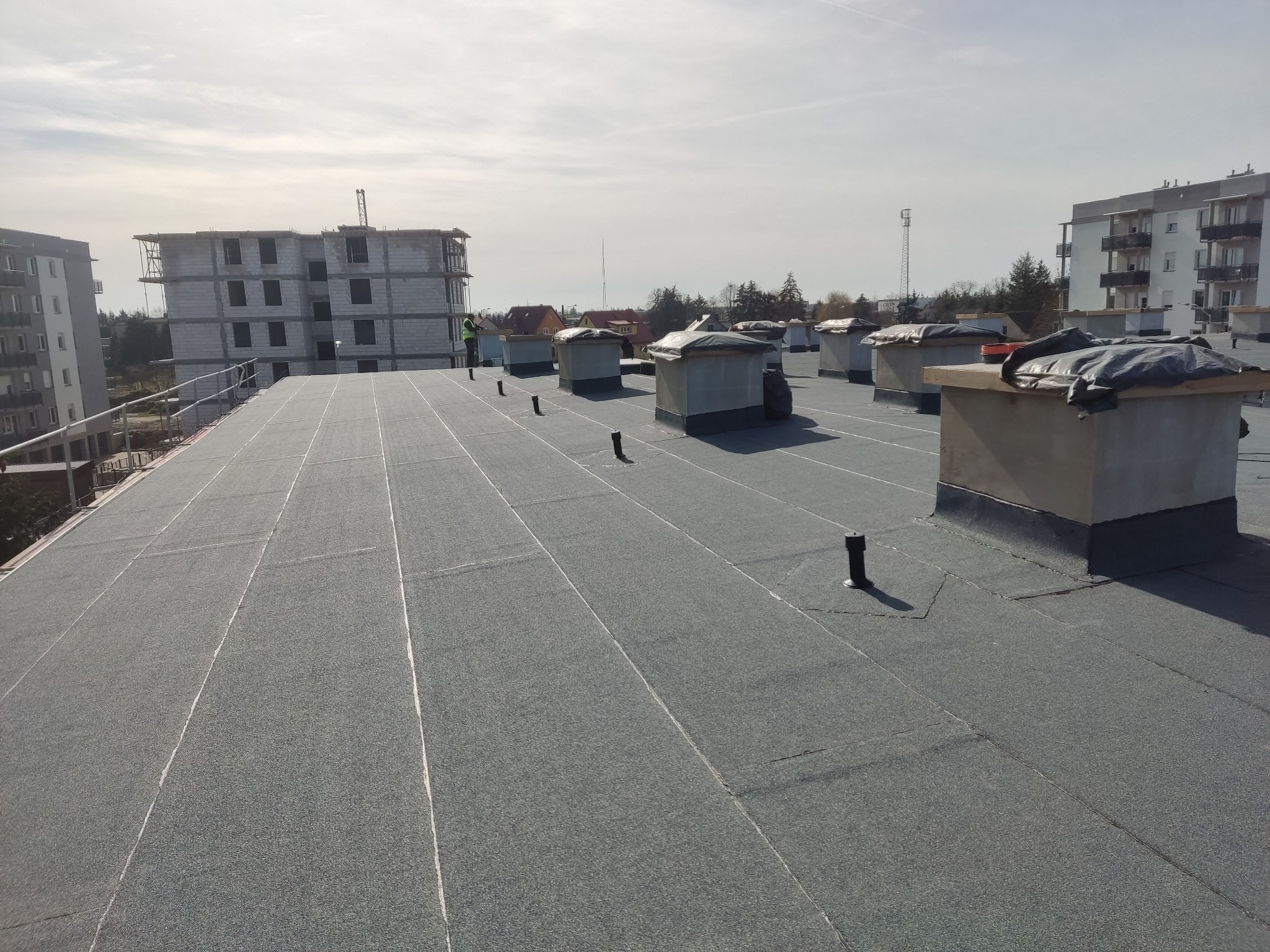 Wykończony płaski dach pokryty papą termozgrzewalną w Frankfurt am Main, z widocznymi kominami wentylacyjnymi, pionami kanalizacyjnymi i pracownikiem w oddali.