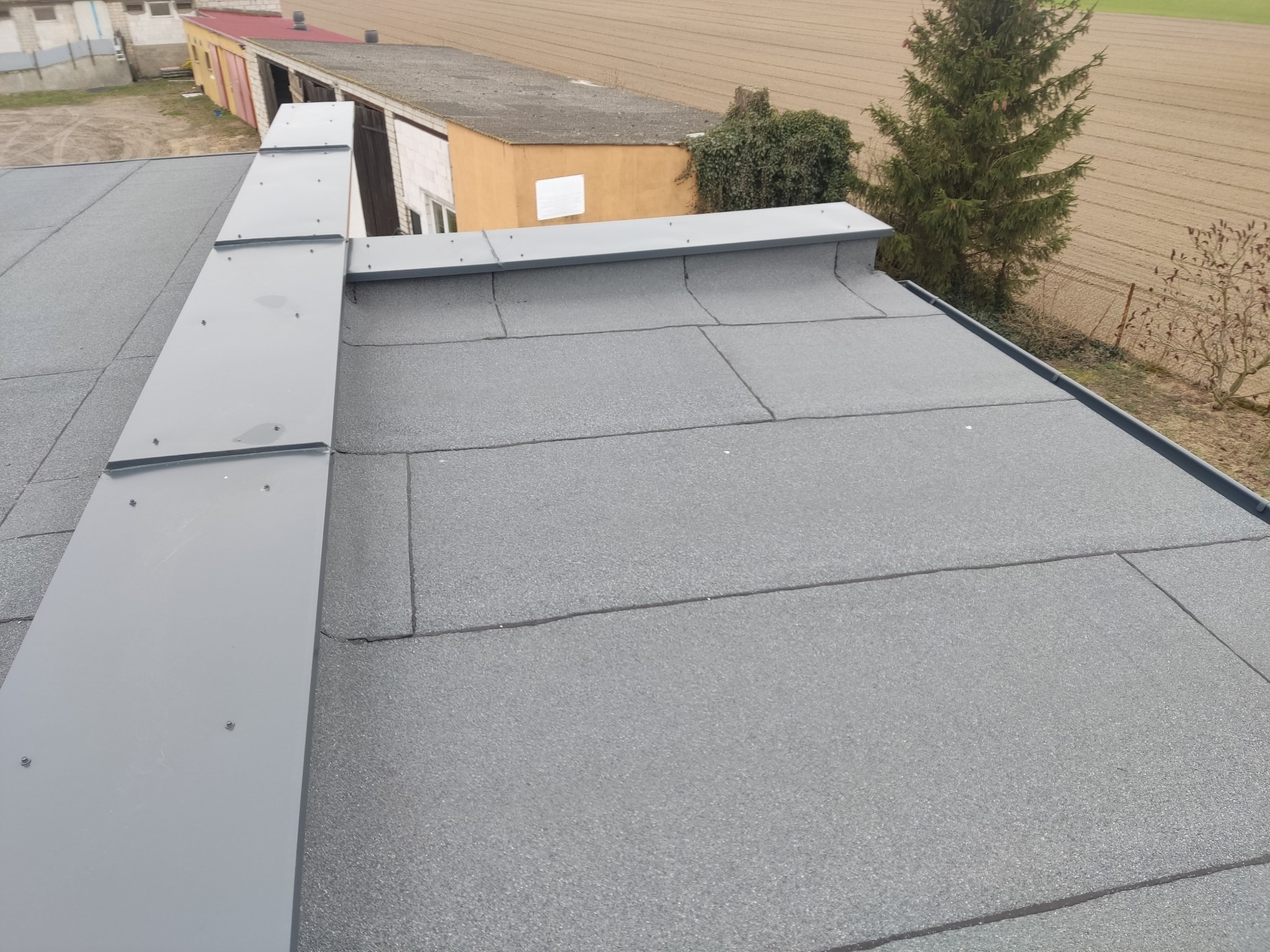 Szary, płaski dach pokryty papą z widocznymi łączeniami, obróbki blacharskie w kolorze szarym, w tle budynki gospodarcze i pole uprawne.