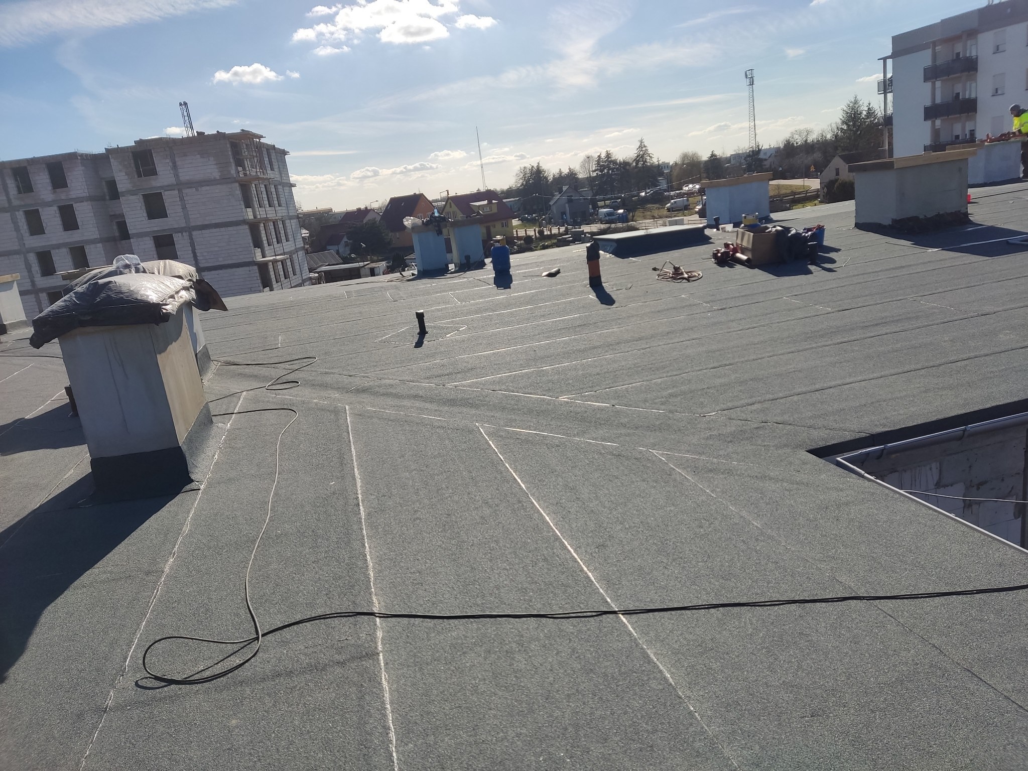 Nowo pokryty, płaski dach budynku wielorodzinnego z widocznymi kominami, obróbkami i przewodami elektrycznymi, w tle budowa i zabudowa miejska w słoneczny dzień.