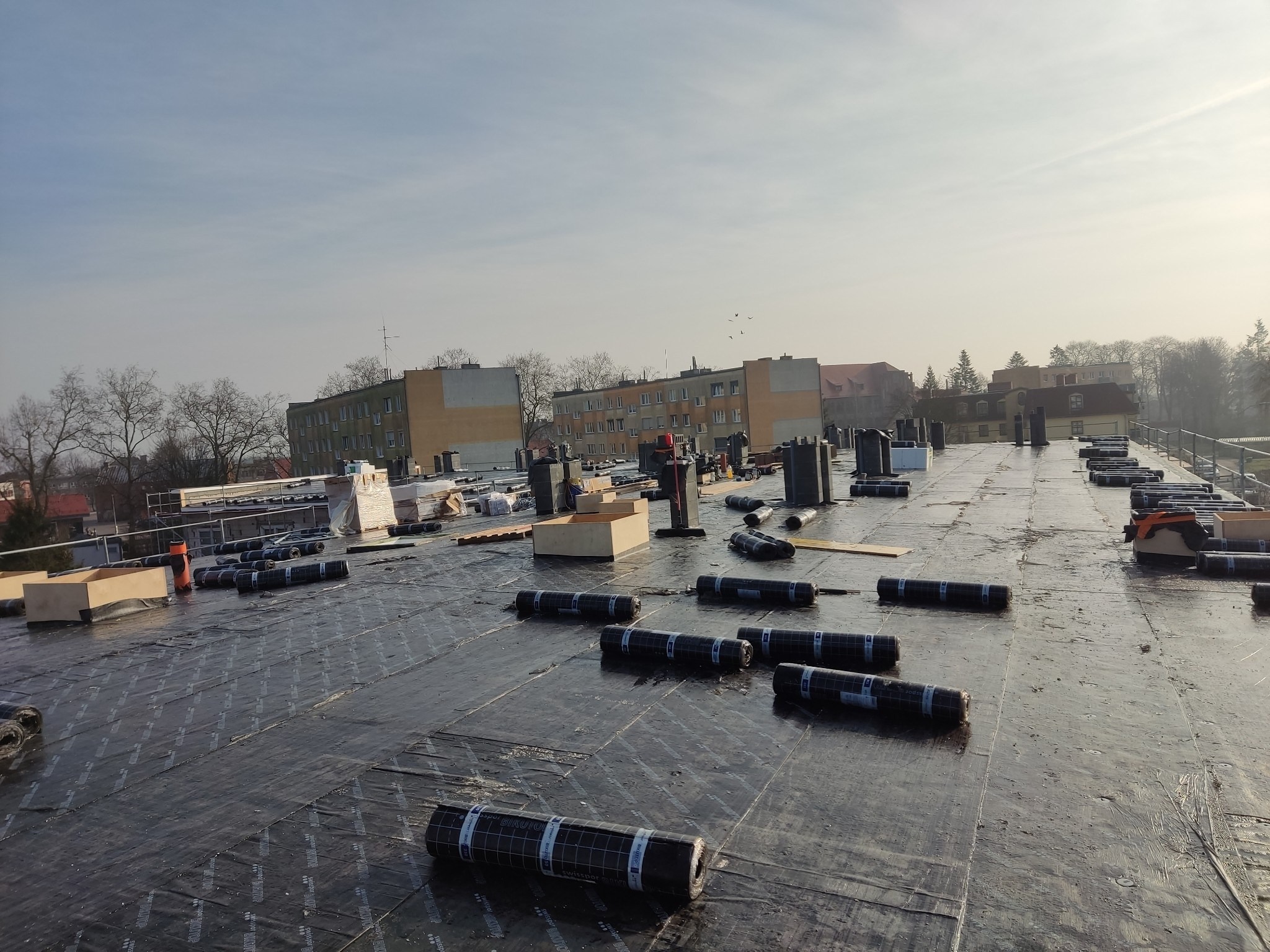 Rozwinięta papa termozgrzewalna na dachu budynku wielorodzinnego w trakcie krycia, widoczne rolki papy, elementy wentylacyjne i narzędzia dekarskie, w tle zabudowa miejska i pochmurne niebo.