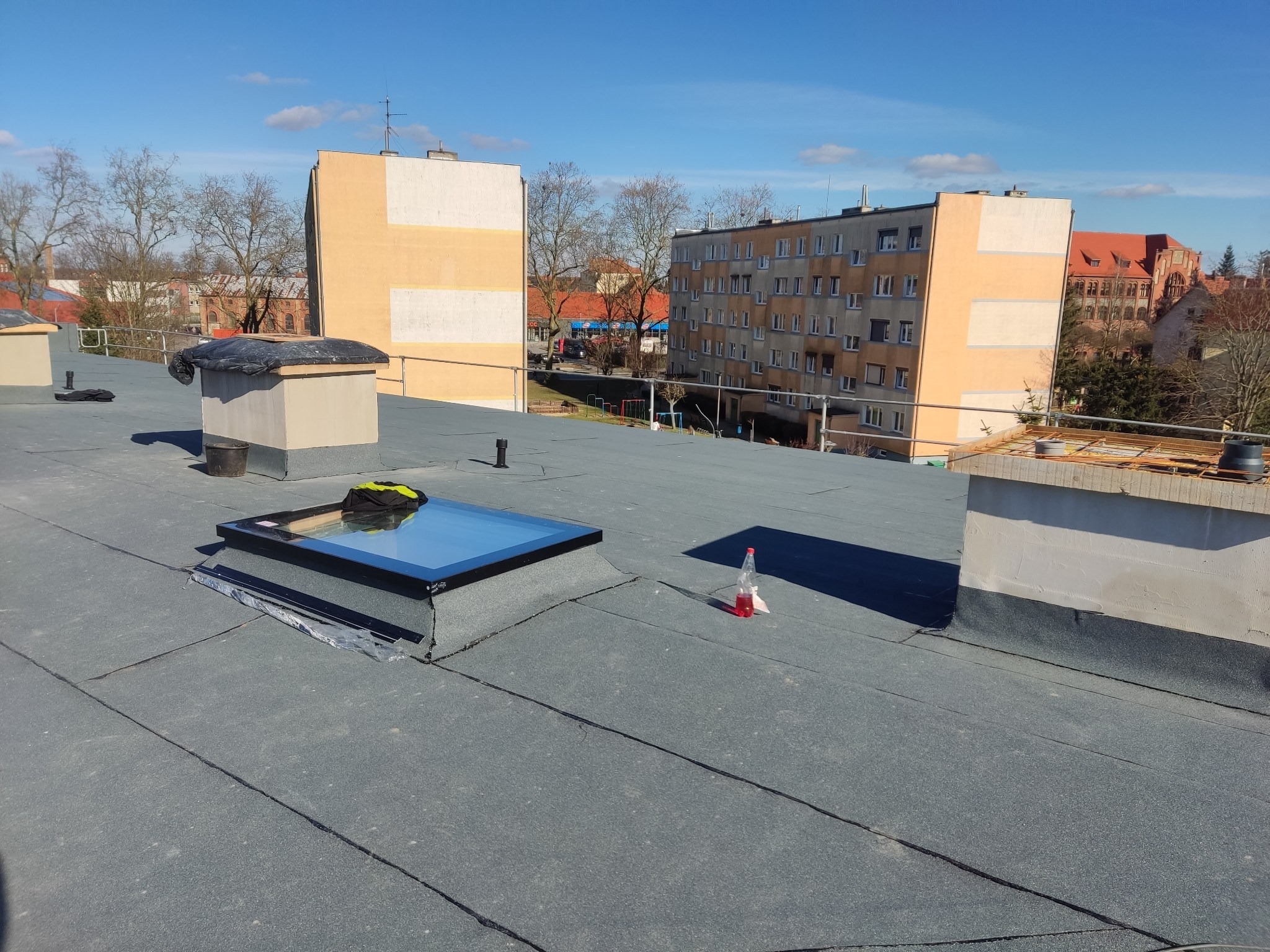 Płaski dach pokryty papą z zamontowanym oknem dachowym; widoczne kominy, butelka z płynem i budynki w tle.