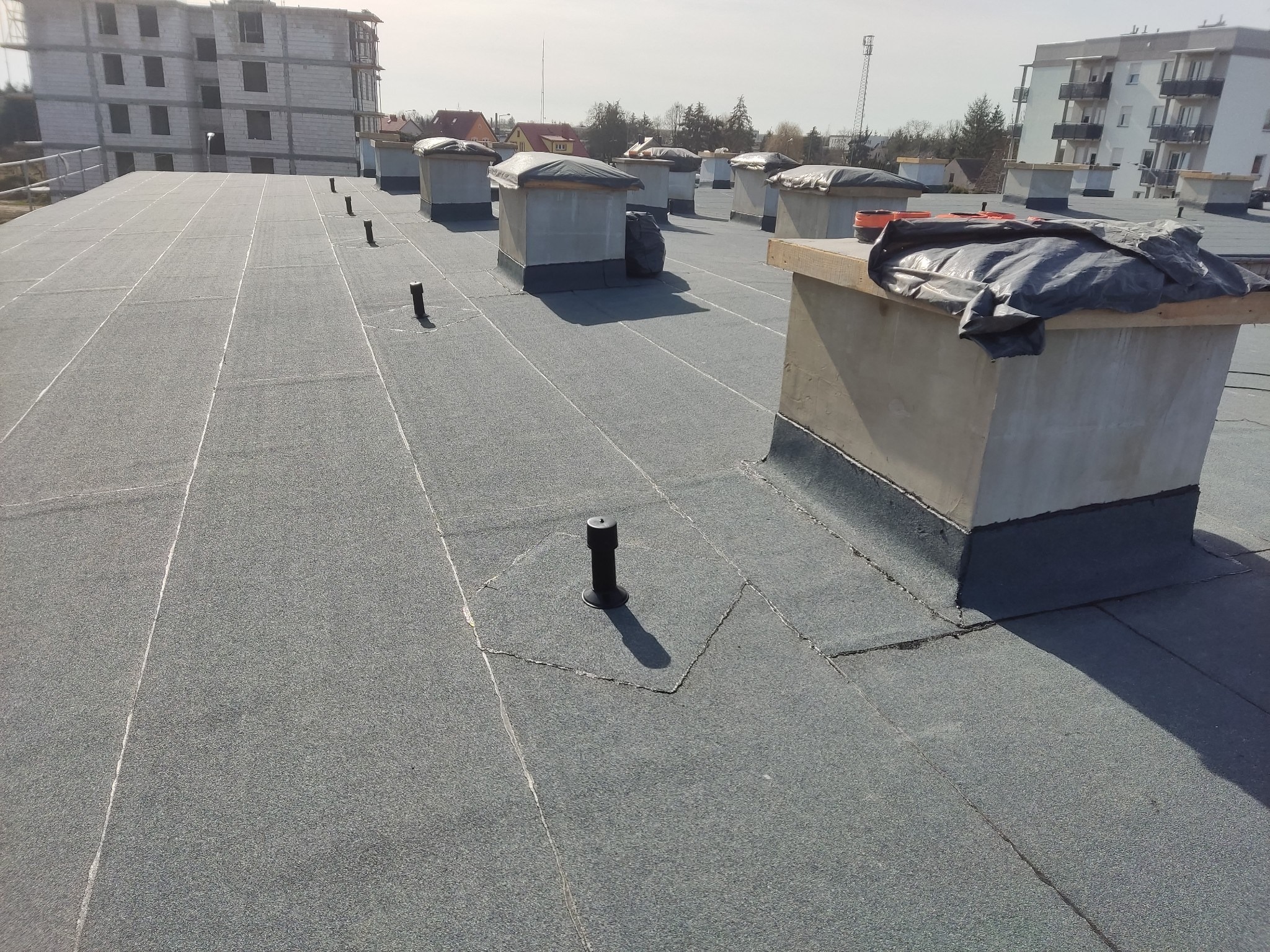 Szary, płaski dach pokryty papą termozgrzewalną z widocznymi kominami zabezpieczonymi folią i elementami wentylacyjnymi na tle budynków mieszkalnych w trakcie budowy.