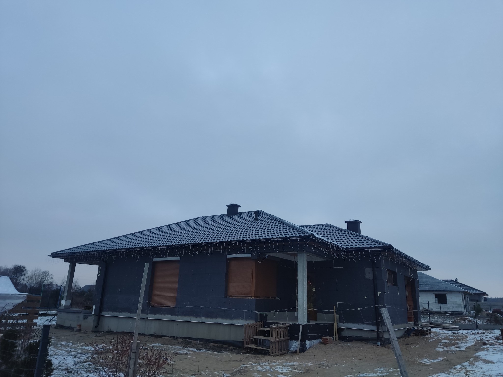 Nowy dom w trakcie budowy z ciemną elewacją i grafitowym dachem pokrytym śniegiem, oświetlony świątecznymi lampkami, na tle pochmurnego nieba.