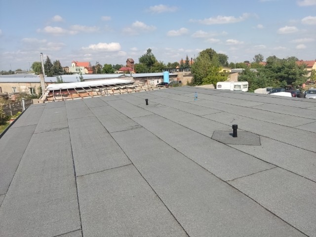 Płaski dach pokryty papą termozgrzewalną w trakcie prac, widoczne kominki wentylacyjne i fragment konstrukcji dachu sąsiedniego budynku w tle.