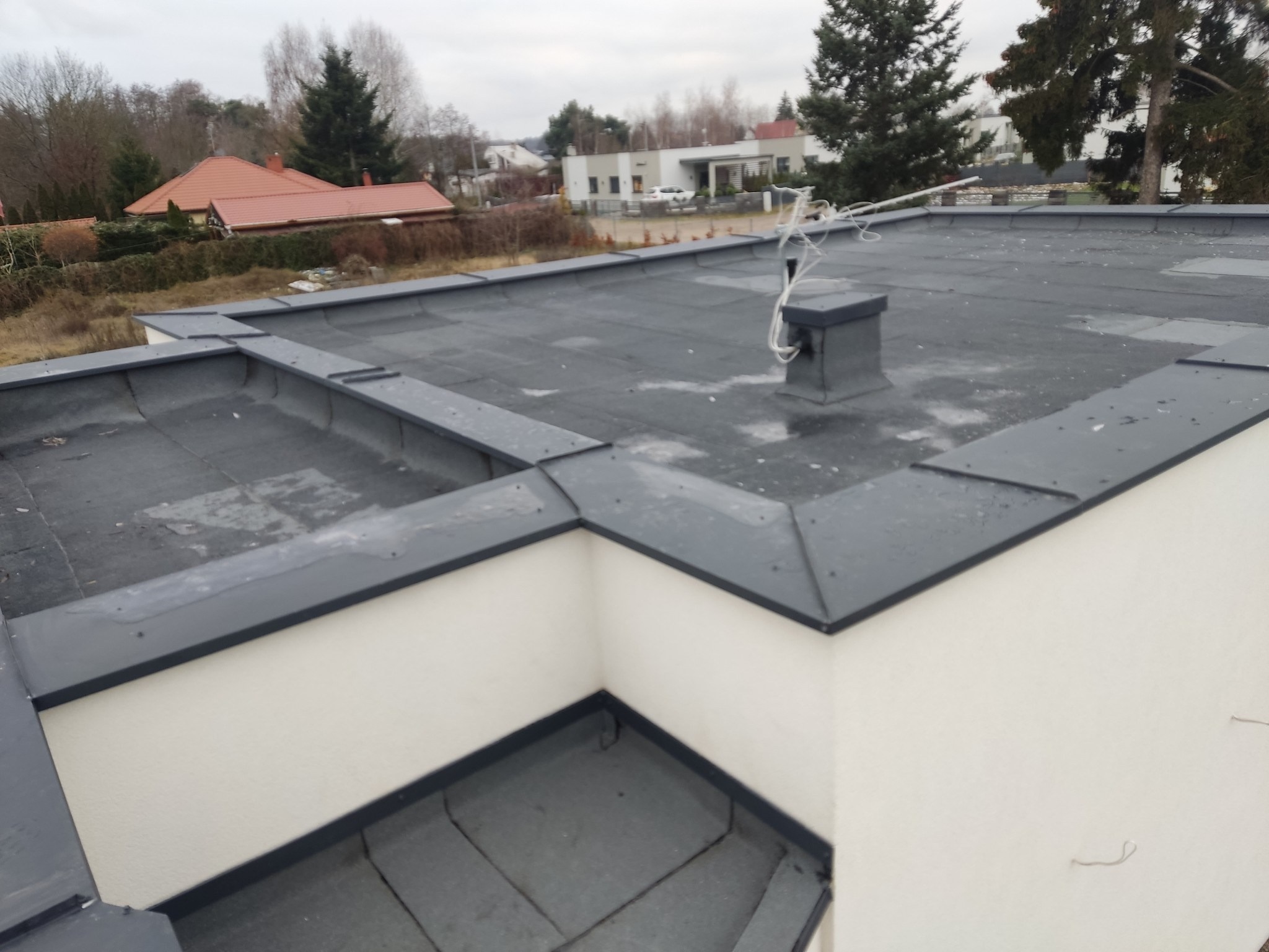 Płaski dach pokryty czarną papą termozgrzewalną z obróbkami blacharskimi w kolorze grafitowym, widoczny komin i instalacja odgromowa, w tle zabudowa jednorodzinna.