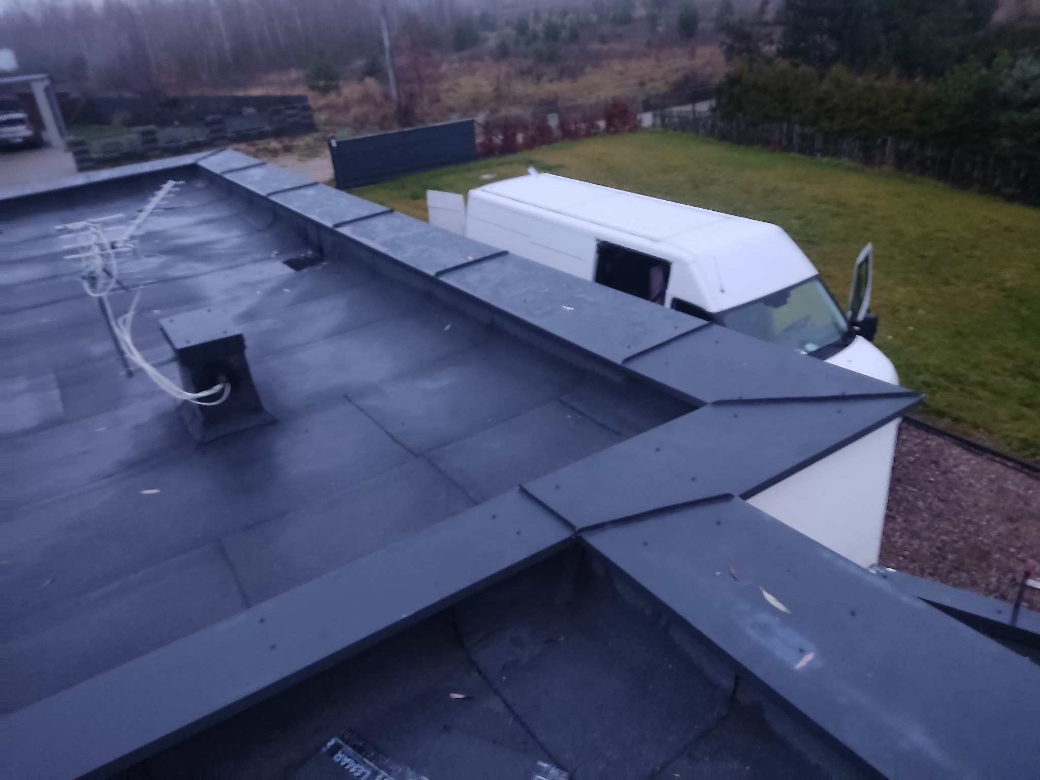 Czarny, płaski dach pokryty papą termozgrzewalną z obróbkami blacharskimi w kolorze grafitowym, antena na dachu, biały bus firmy dekarskiej w tle.