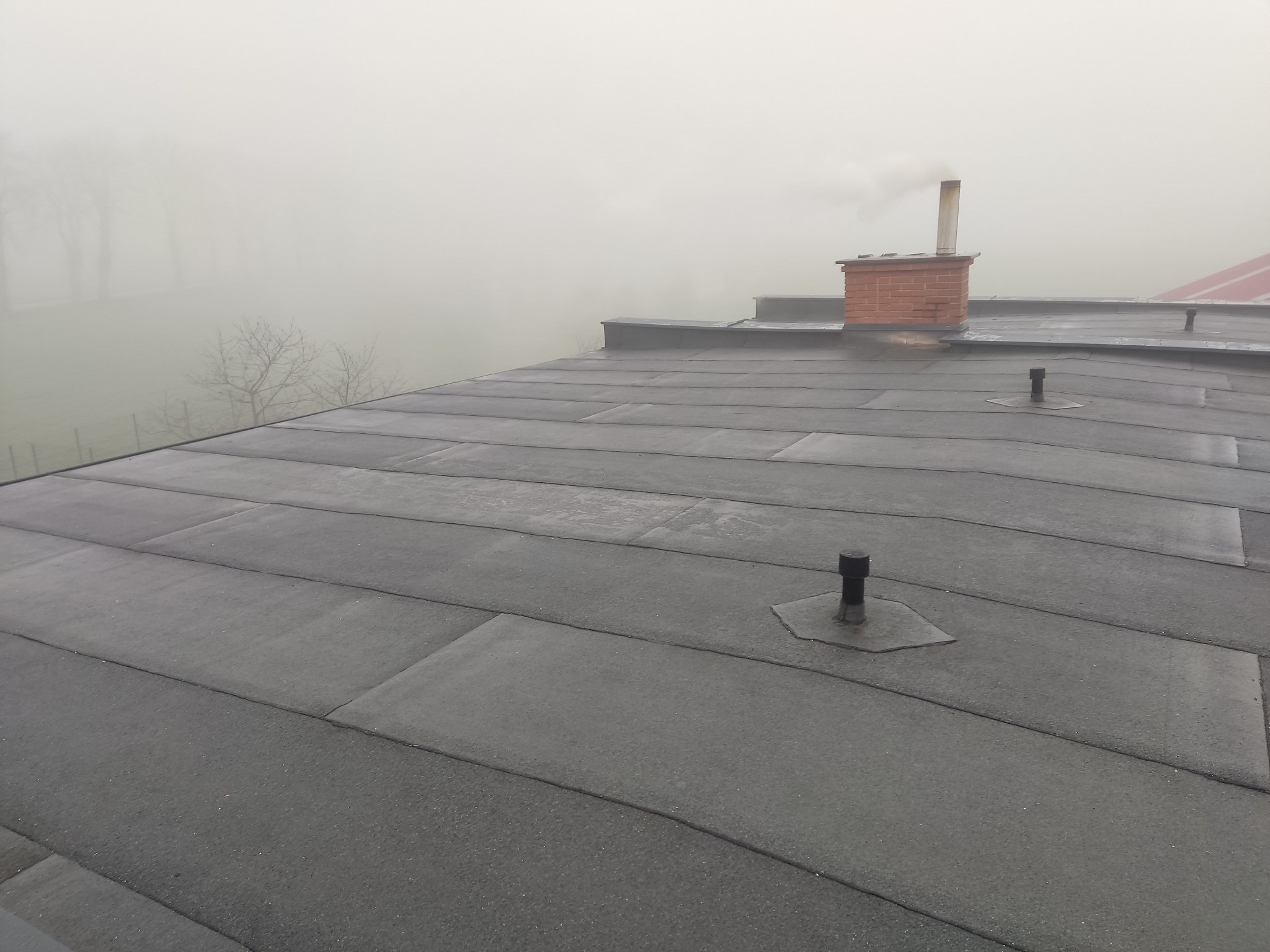 Widok z góry na dach pokryty papą bitumiczną w kształcie prostokątnych płyt, z widocznym kominem z czerwonej cegły, odpowietrznikami i mglistym krajobrazem w tle.