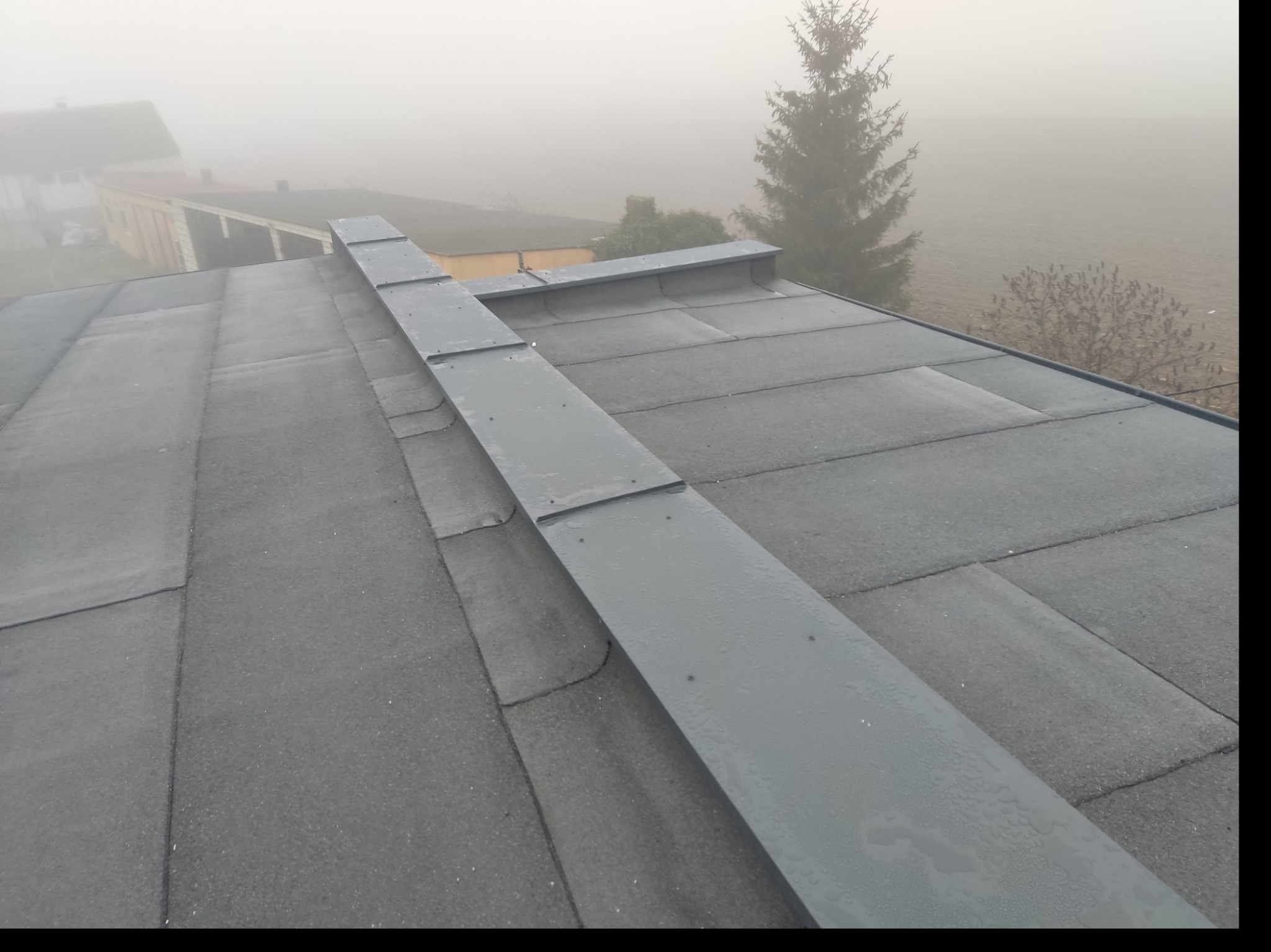 Widok z góry na płaski dach pokryty papą z zamontowaną, szarą, metalową obróbką blacharską w mglisty dzień. W tle widoczne budynki i drzewa.