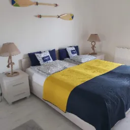 Jasna sypialnia z dwoma łóżkami, ozdobnymi wiosłami na ścianie, lampkami nocnymi z podstawami owiniętymi sznurem i kołdrą w kolorach żółtym i granatowym.