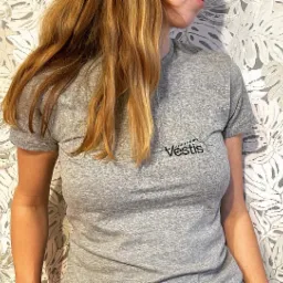 Kobieta w szarej koszulce z czarnym napisem 'Vestis' i zielonym symbolem nici i igły na tle ściany w liście monstery.