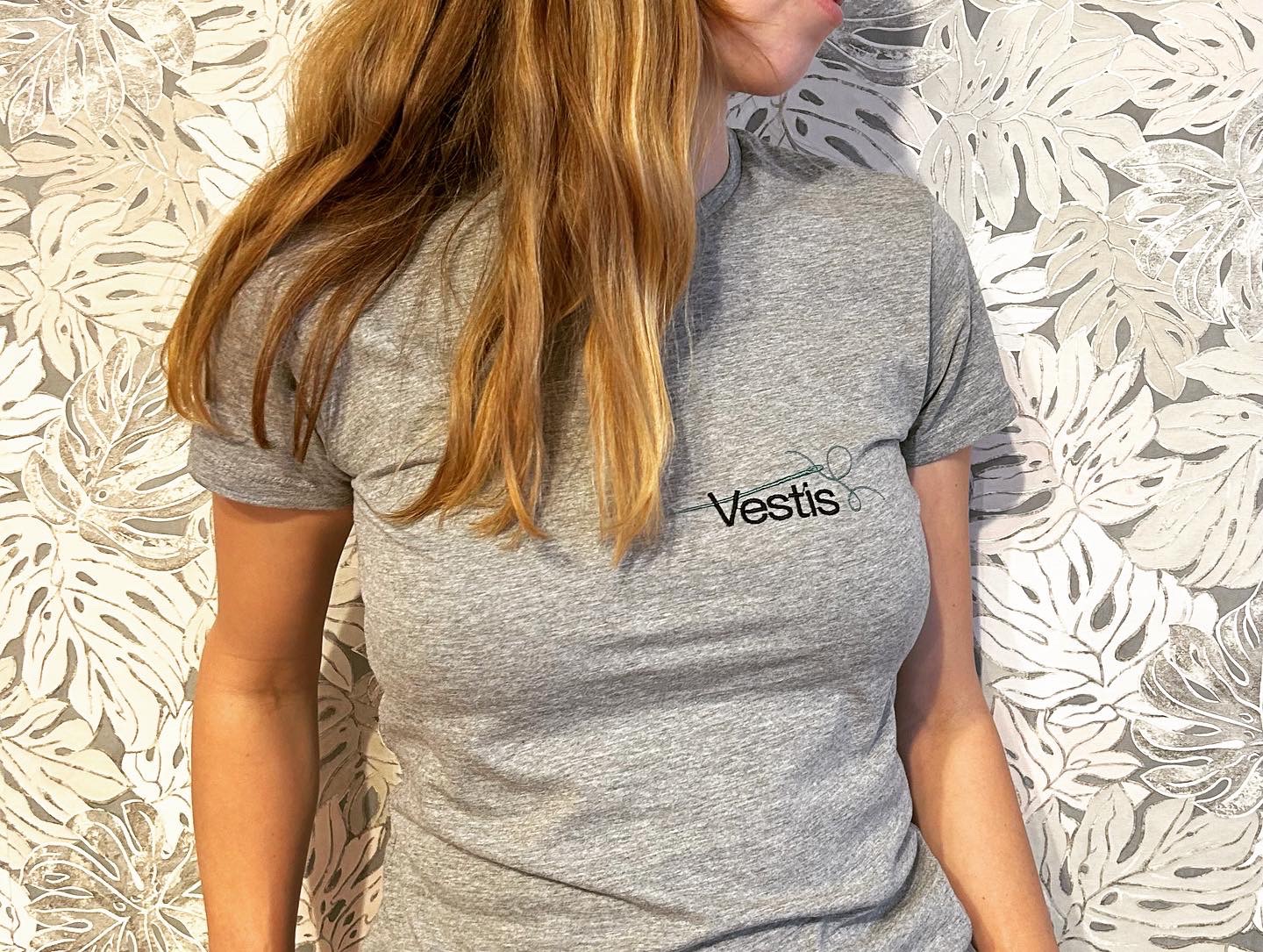 Kobieta w szarej koszulce z czarnym napisem 'Vestis' i zielonym symbolem nici i igły na tle ściany w liście monstery.