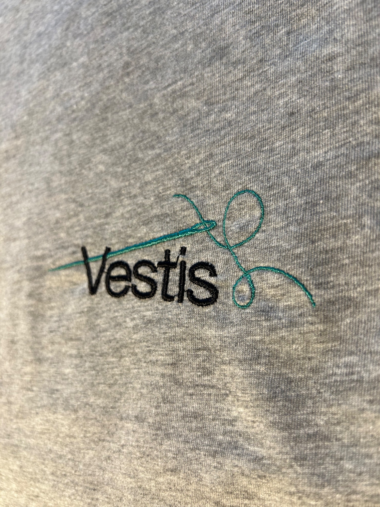 Szara koszulka z wyhaftowanym, turkusowym wzorem igły z nitką i czarnym napisem 'Vestis'.