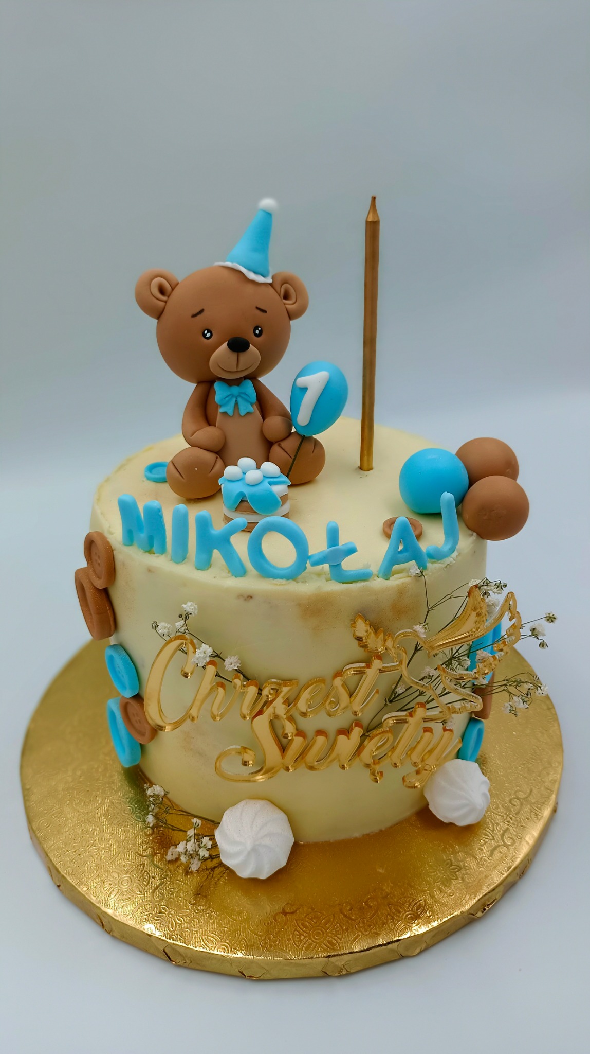 Tort urodzinowy z kremowym lukrem, dekorowany niebieskimi literami układającymi imię 'Mikołaj', złotym napisem 'Chrzest Święty', figurką brązowego misia w niebieskim kapeluszu i z balonem z cyfrą...