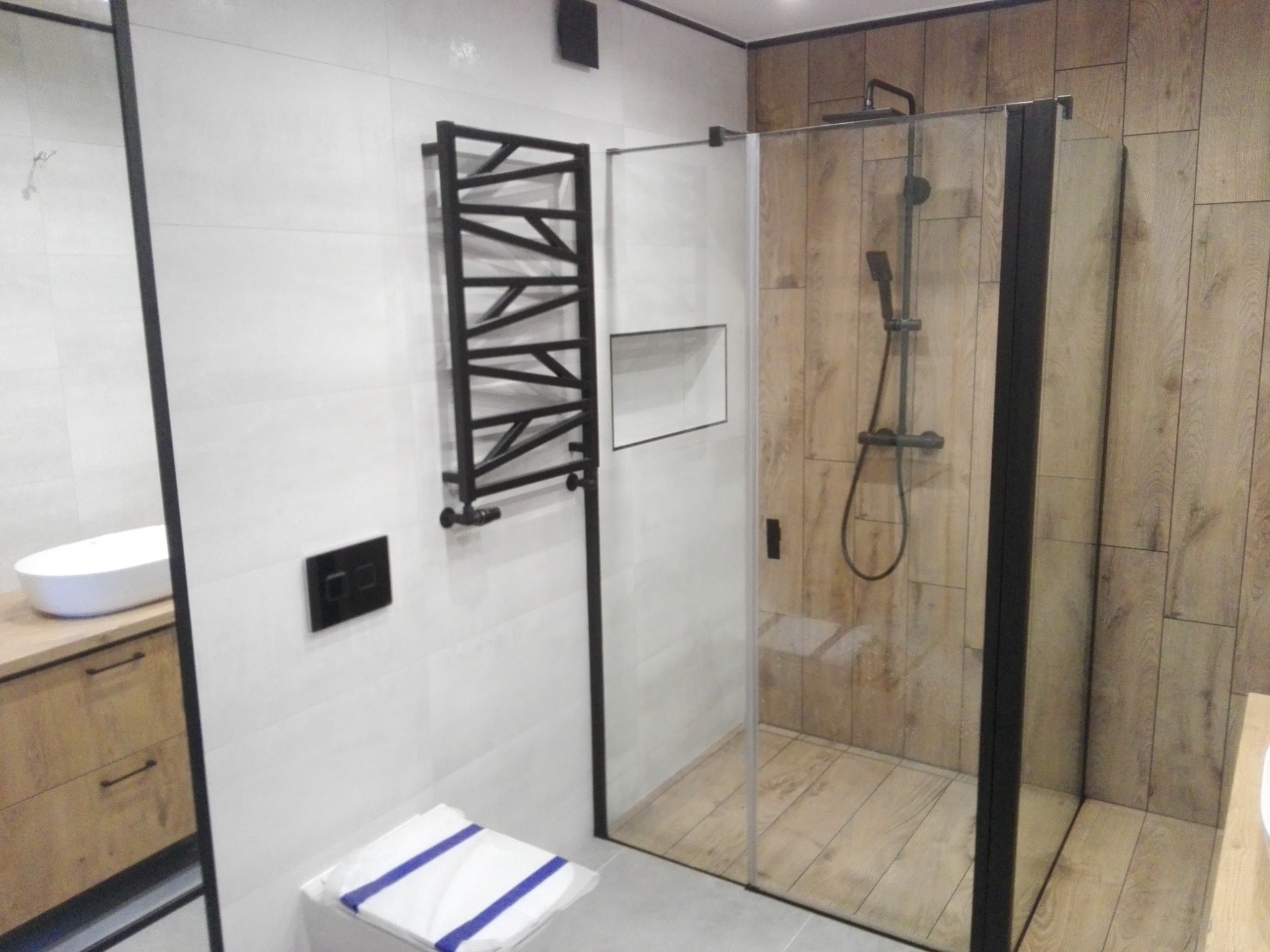 Nowoczesna łazienka z prysznicem typu walk-in z czarnymi ramami, grzejnikiem drabinkowym w geometrycznym stylu i drewnopodobnymi płytkami na ścianach prysznica.