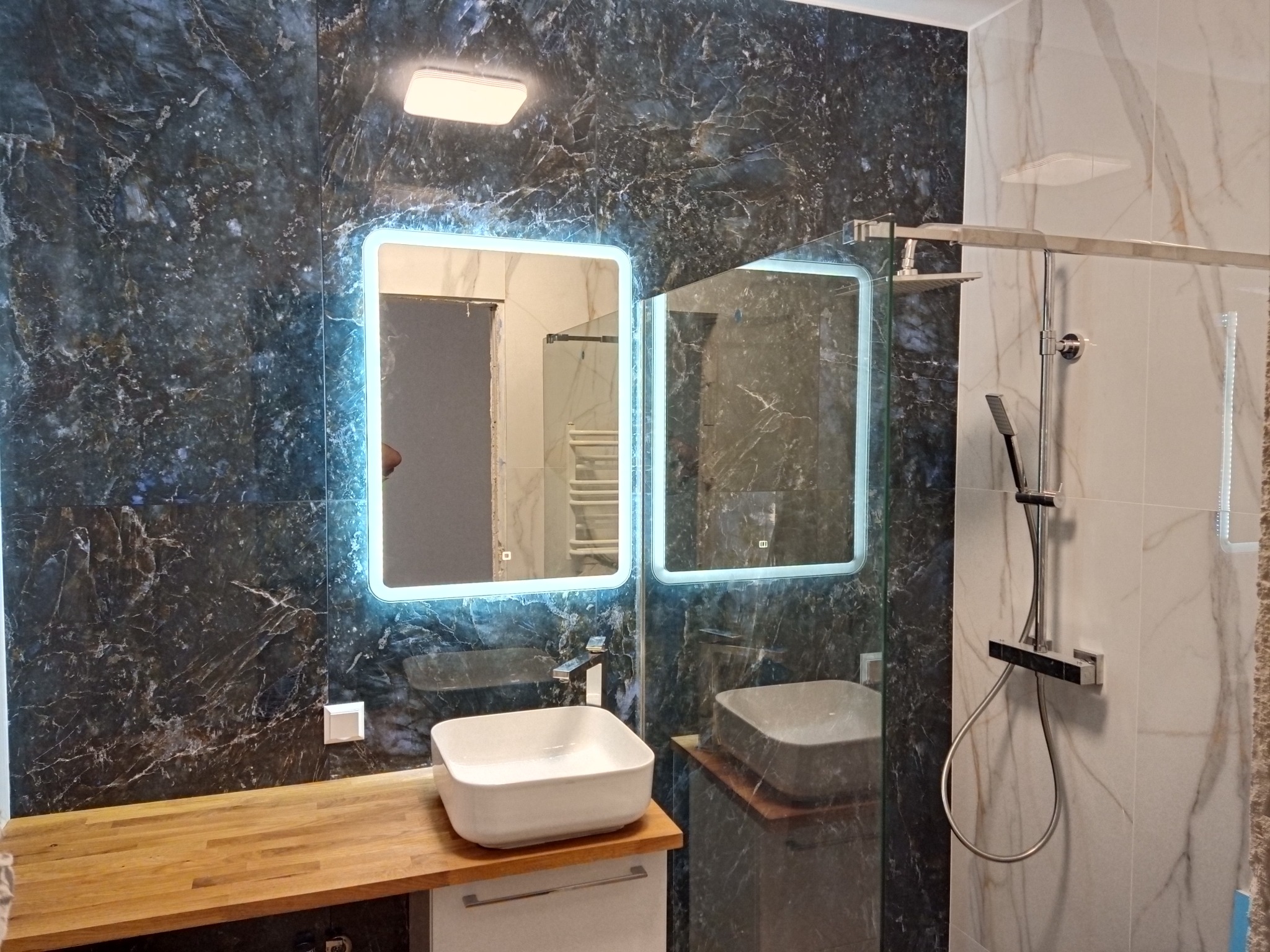 Nowoczesna łazienka z podświetlanym lustrem, umywalką nablatową na drewnianym blacie i kabiną prysznicową ze szklaną ścianką, wykończona płytkami imitującymi marmur.