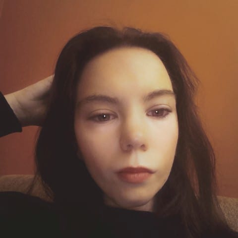 Portret młodej kobiety z ciemnymi włosami, spoglądającej w dół, z ręką uniesioną do włosów na tle pomarańczowej ściany.