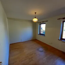 Pusty pokój w Gdańsku przed remontem, z drewnianą podłogą, grzejnikiem pod oknem i lampą wiszącą na suficie, ściany przygotowane do malowania.