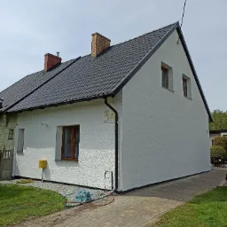 Dom jednorodzinny z nową, białą elewacją o strukturze baranka, ciemnym dachem i numerem '34' na ścianie. Widoczne narzędzia i przewody sugerują trwające prace.