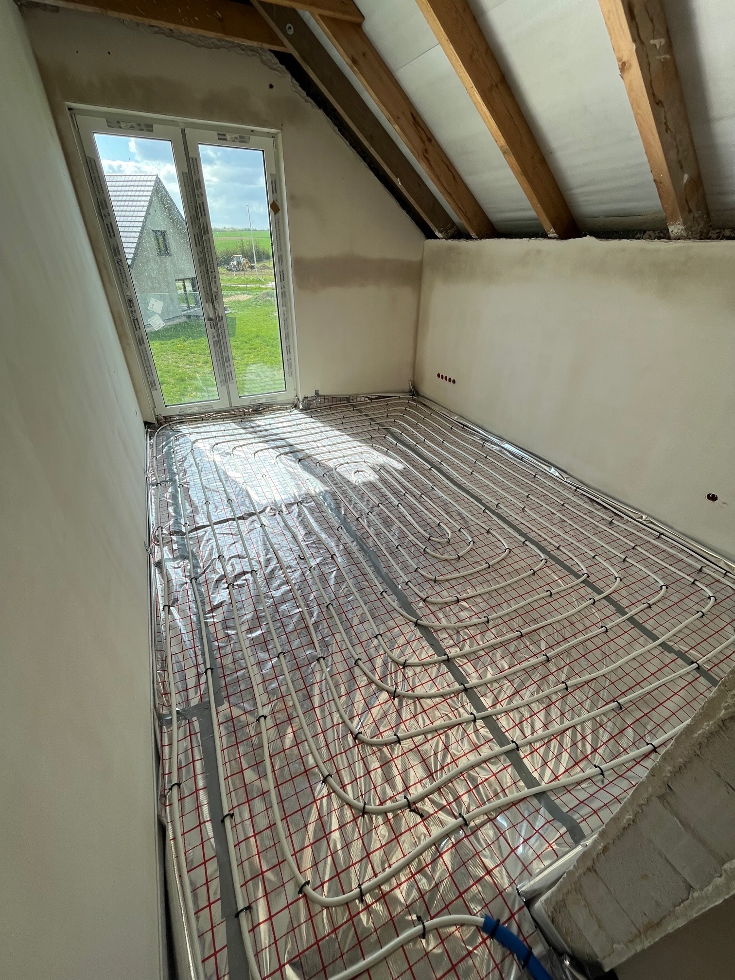 Instalacja ogrzewania podłogowego w poddaszowym pokoju z widokiem na dom i pole przez okno. Rury grzewcze ułożone na folii z czerwoną siatką, mocowane czarnymi klipsami. Widoczne drewniane krokwie...